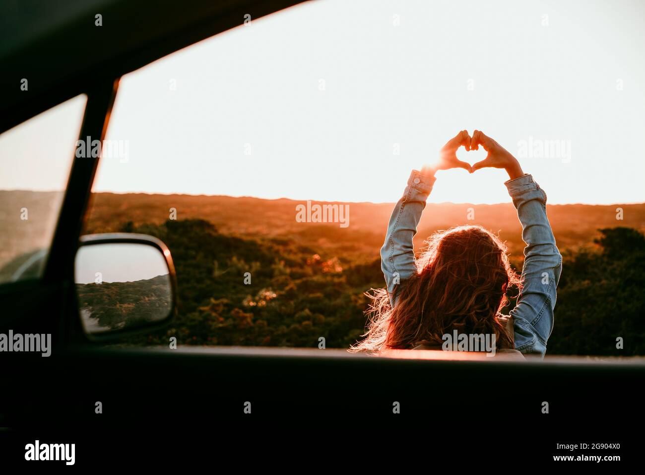 Donna che fa la forma del cuore con le mani viste attraverso il finestrino dell'automobile durante il tramonto Foto Stock