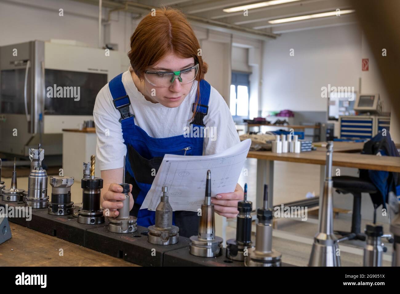 Tecnico femminile che lavora con parti di macchinari metallici durante la lettura del libro in officina Foto Stock
