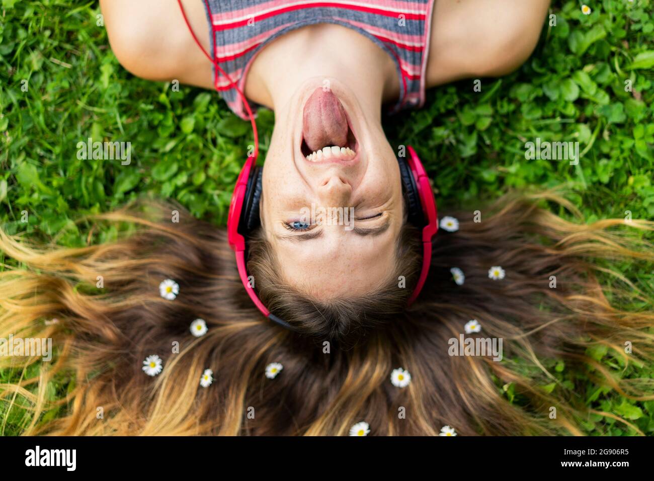 Giovane donna maliziosa che sporge lingua con fiori in capelli giacenti sull'erba Foto Stock