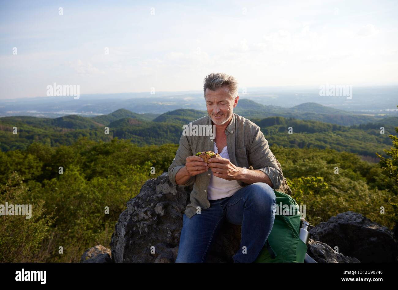 Uomo sorridente che mangia cibo mentre si siede in cima alla montagna Foto Stock