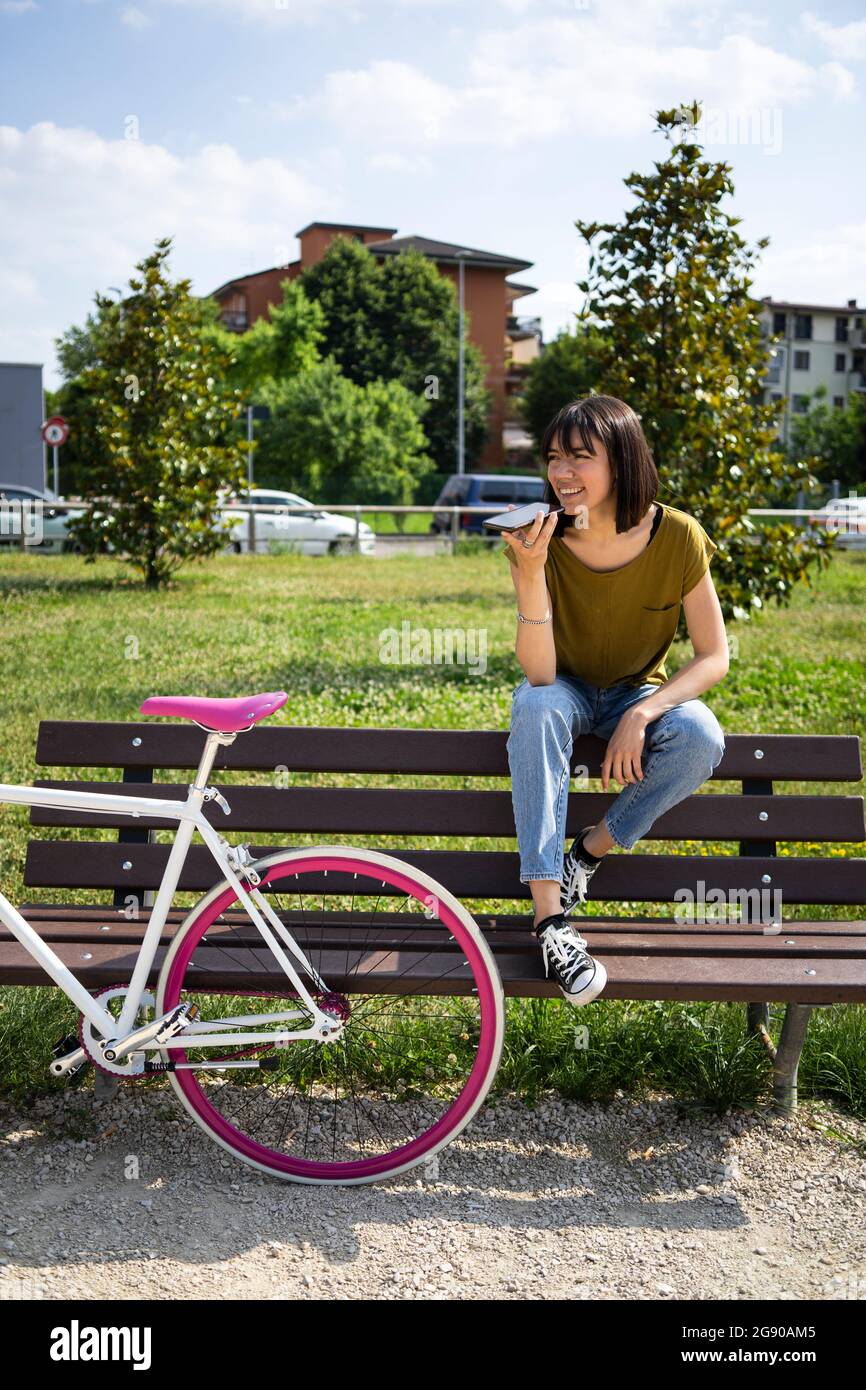 Giovane donna in bicicletta che invia messaggi vocali tramite smartphone mentre si siede sulla panchina del parco Foto Stock
