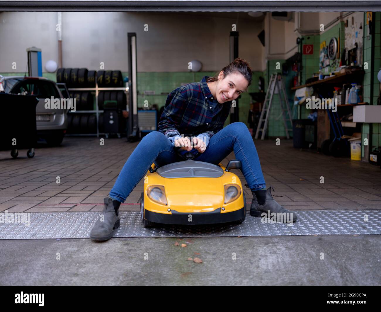 Meccanico femminile sorridente seduto su un'auto giocattolo presso il negozio di riparazione auto Foto Stock