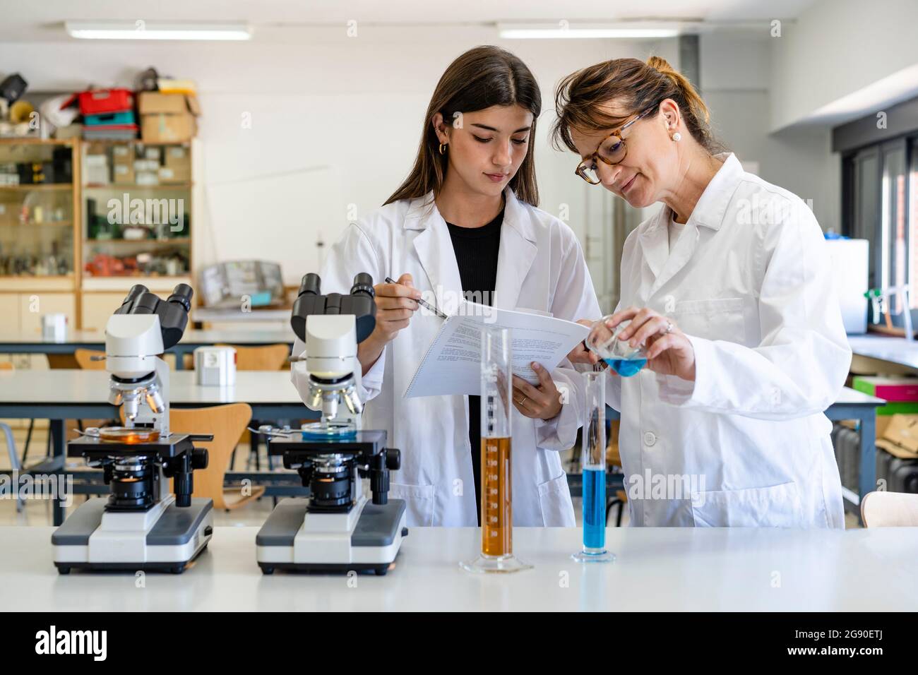Scienziata femminile che legge il documento mentre il collega versa il liquido in un cilindro graduato Foto Stock