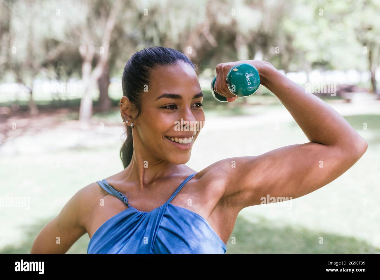 Atleta femminile sorridente mentre tiene il dumbbell e i muscoli flettenti al parco Foto Stock