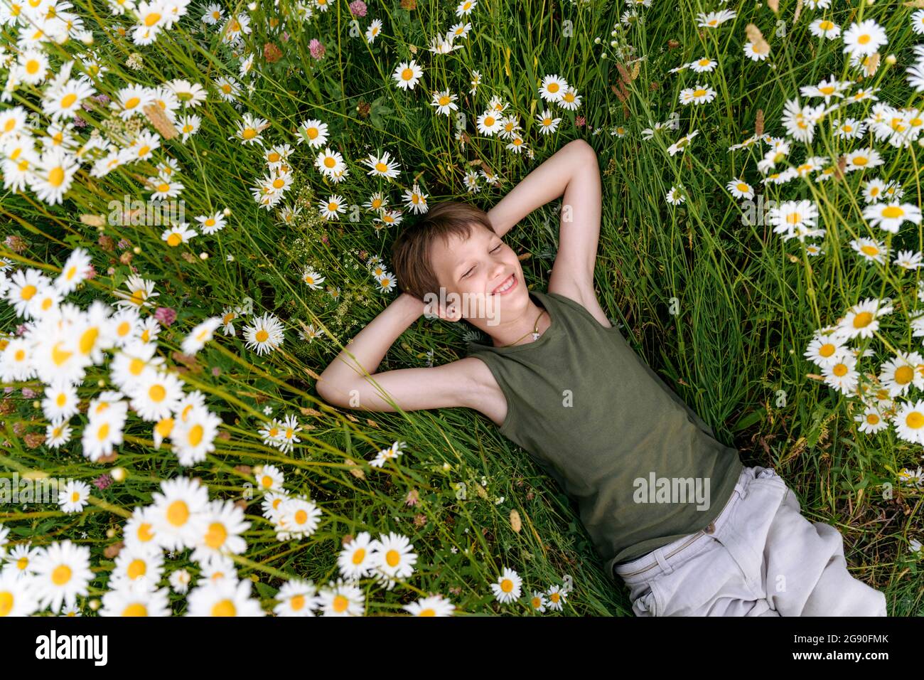 Ragazzo sorridente con le mani dietro la testa adagiato su piante camomilla in campo Foto Stock