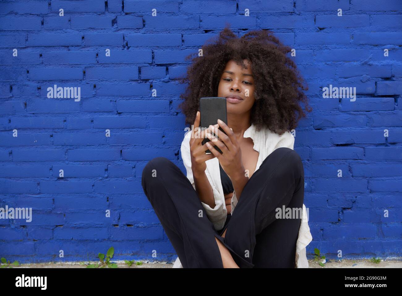 Giovane donna afrofroica che naviga in rete attraverso il cellulare di fronte al muro blu Foto Stock