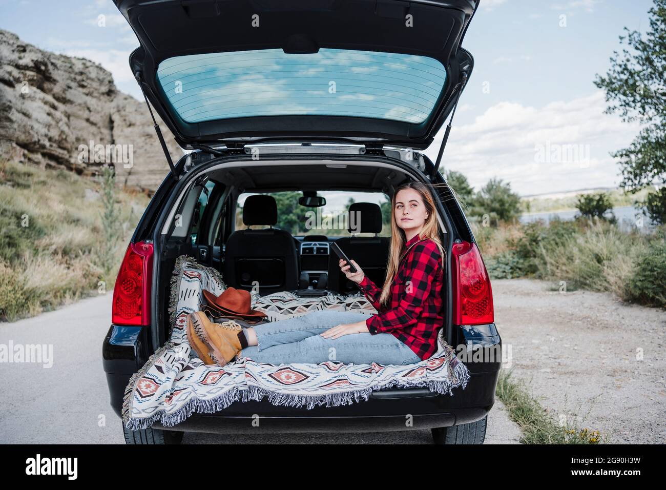 Giovane donna bionda seduta con smartphone nel bagagliaio dell'auto Foto Stock