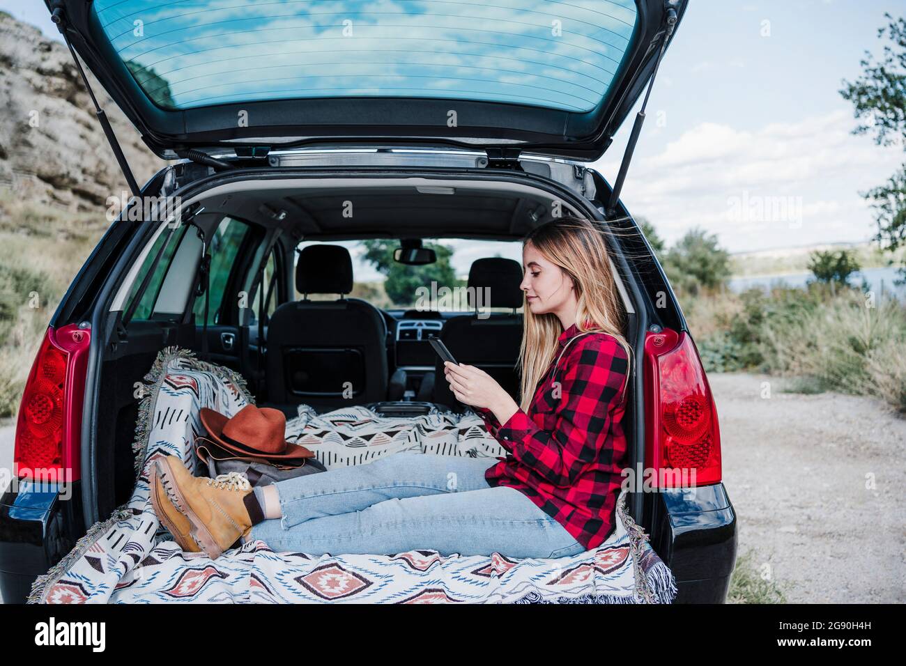 Giovane donna che invia messaggi di testo tramite smartphone mentre si siede nel bagagliaio dell'auto Foto Stock