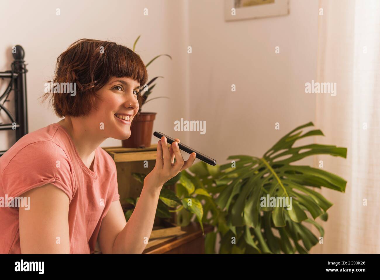 Donna sorridente che invia messaggi vocali tramite smartphone seduto in camera da letto a casa Foto Stock