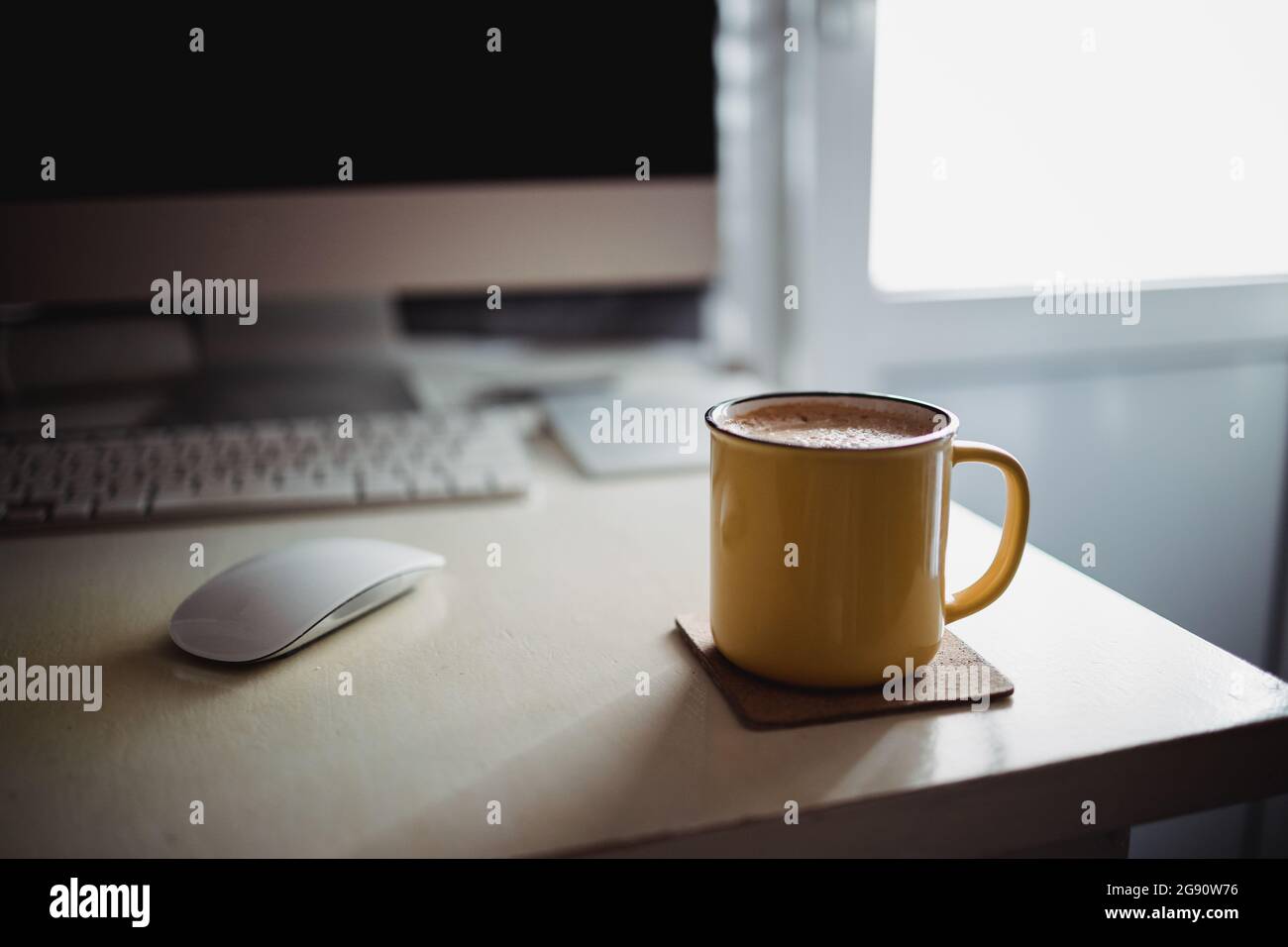 Una tazza di caffè gialla si trova sulla scrivania dell'ufficio, nella soleggiata luce del mattino-sera, accanto al monitor e alla tastiera. Foto Stock