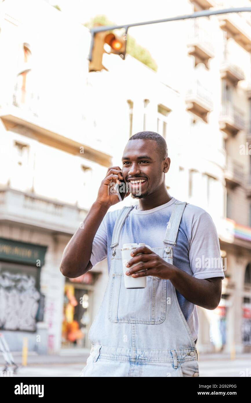 allegro uomo afro sorridente e che parla al telefono. Sta vestendo i vestiti estivi casual e tiene una tazza riutilizzabile di caffè Foto Stock