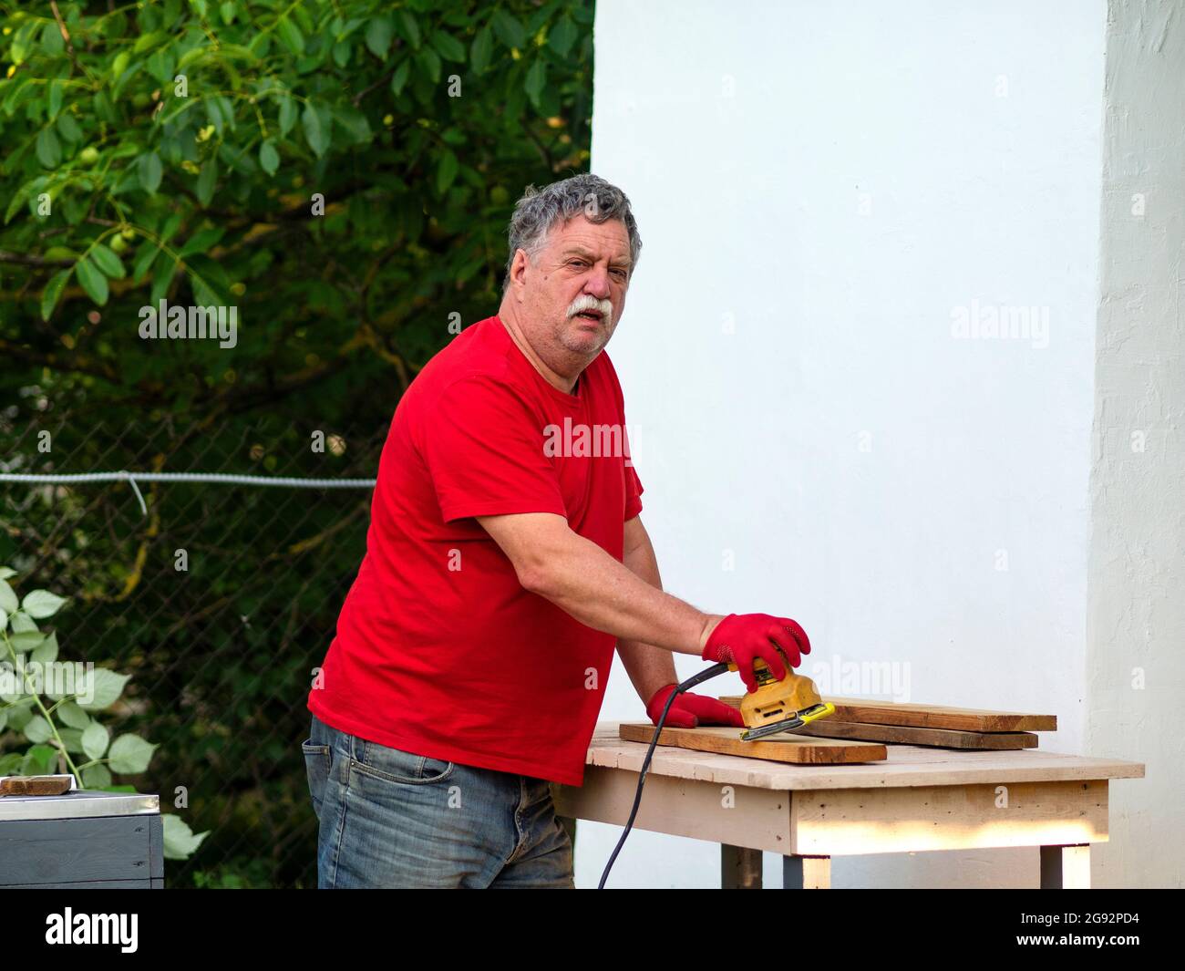 L'uomo caucasico anziano con un baffi in una t-shirt rossa sta levigando un bordo usando una levigatrice su una disposizione in un giardino vicino ad una parete bianca di una casa Foto Stock