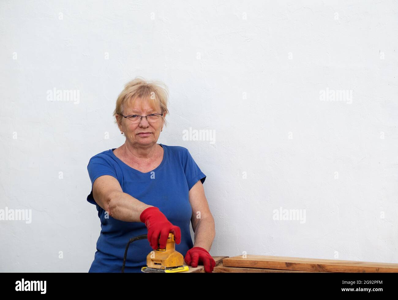 Donna caucasica anziana in una t-shirt blu e guanti rossi sta levigando una tavola con una levigatrice su un layout in un giardino vicino a una parete bianca a casa Foto Stock