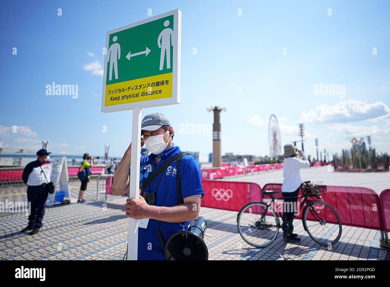 Tokio, Giappone. 24 luglio 2021. I visitatori guardano alla fiamma olimpica. Un membro dello staff ha un cartello con il simbolo e la scritta "Mantieni la distanza fisica" per mantenere una distanza sufficiente a proteggere se stesso e gli altri dal contrarre il coronavirus. Le Olimpiadi di Tokyo 2020 si terranno dal 23 luglio 2021 all'8 agosto 2021. Credit: Michael Kappeler/dpa/Alamy Live News Foto Stock