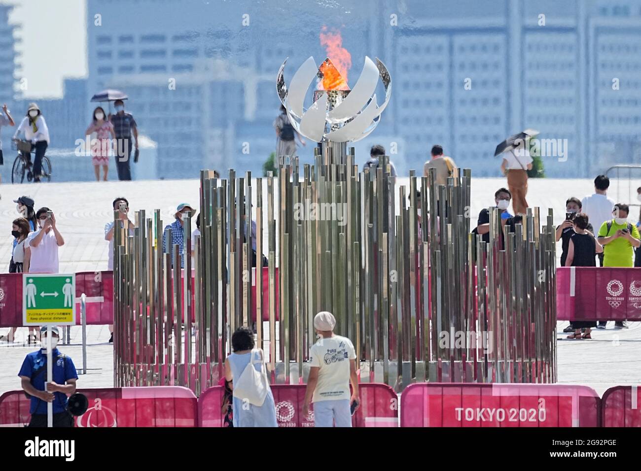 Tokio, Giappone. 24 luglio 2021. I visitatori guardano alla fiamma olimpica. Un membro del personale ha un cartello con il simbolo e la dicitura "Mantieni la distanza fisica" per mantenere una distanza sufficiente a proteggere se stesso e gli altri dal contrarre il coronavirus SARS-COV-2. Le Olimpiadi di Tokyo 2020 si terranno dal 23 luglio 2021 all'8 agosto 2021. Credit: Michael Kappeler/dpa/Alamy Live News Foto Stock