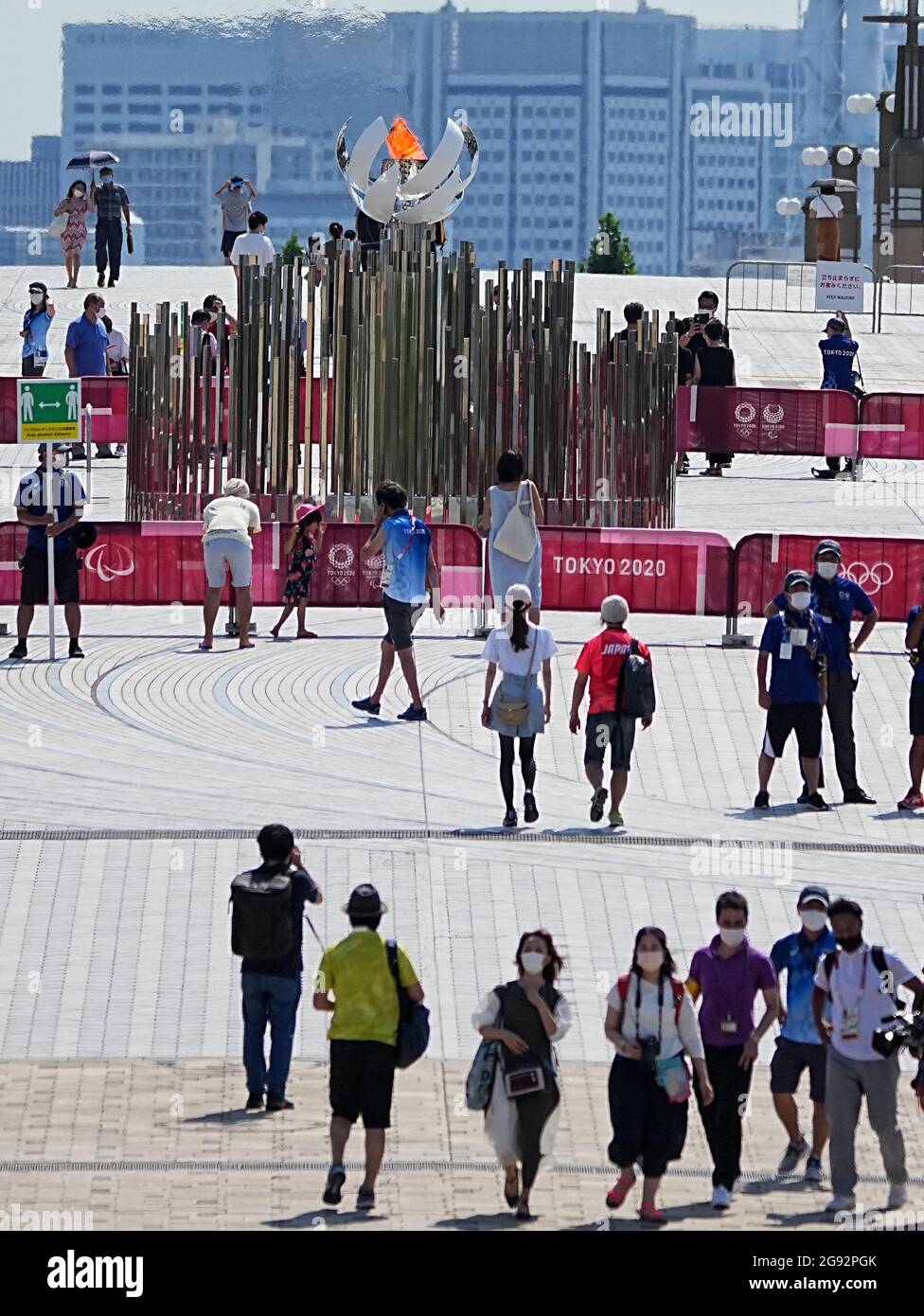 Tokio, Giappone. 24 luglio 2021. I visitatori guardano alla fiamma olimpica. Un membro del personale ha un cartello con il simbolo e la dicitura "Mantieni la distanza fisica" per mantenere una distanza sufficiente a proteggere se stesso e gli altri dal contrarre il coronavirus SARS-COV-2. Le Olimpiadi di Tokyo 2020 si terranno dal 23 luglio 2021 all'8 agosto 2021. Credit: Michael Kappeler/dpa/Alamy Live News Foto Stock