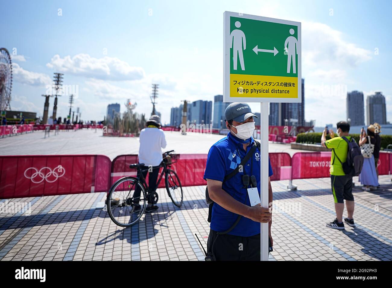 Tokio, Giappone. 24 luglio 2021. I visitatori guardano alla fiamma olimpica. Un membro dello staff ha un cartello con il simbolo e la scritta "Mantieni la distanza fisica" per mantenere una distanza sufficiente a proteggere se stesso e gli altri dal contrarre il coronavirus. Le Olimpiadi di Tokyo 2020 si terranno dal 23 luglio 2021 all'8 agosto 2021. Credit: Michael Kappeler/dpa/Alamy Live News Foto Stock