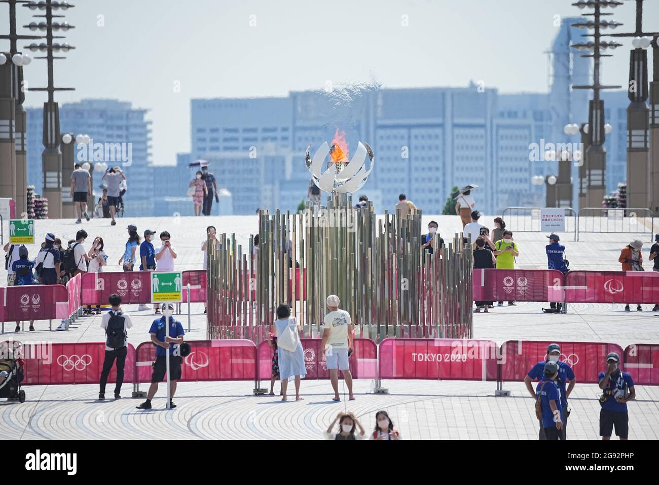 Tokio, Giappone. 24 luglio 2021. I visitatori guardano alla fiamma olimpica. Un membro del personale ha un cartello con il simbolo e la dicitura "Mantieni la distanza fisica" per mantenere una distanza sufficiente a proteggere se stesso e gli altri dal contrarre il coronavirus SARS-COV-2. Le Olimpiadi di Tokyo 2020 si terranno dal 23 luglio 2021 all'8 agosto 2021. Credit: Michael Kappeler/dpa/Alamy Live News Foto Stock