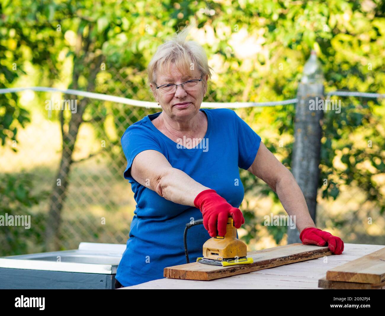 Donna caucasica anziana in una t-shirt blu e guanti rossi sta levigando una tavola con una levigatrice su un layout in un giardino Foto Stock