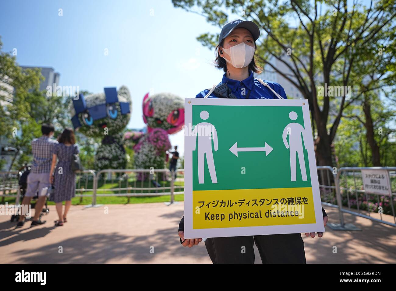 Tokio, Giappone. 24 luglio 2021. I visitatori guardano i personaggi Miraitowa (l), mascotte ufficiale delle Olimpiadi estive di Tokyo 2020, e Someity, mascotte ufficiale delle Paralimpiadi. Un membro dello staff tiene un cartello con un simbolo e le parole 'Keep Physical Distance' per mantenere una distanza sufficiente a proteggere se stessi e gli altri dal contrarre il coronavirus. Le Olimpiadi di Tokyo 2020 si terranno dal 23 luglio 2021 all'8 agosto 2021. Credit: Michael Kappeler/dpa/Alamy Live News Foto Stock