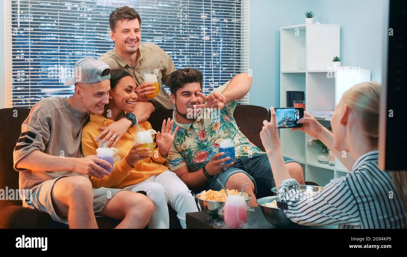 Primo piano della ragazza facendo foto di amici seduti sul divano con cocktail da smartphone. Il gruppo di persone allieterono, sorridendo e posando sulla fotocamera. Foto Stock