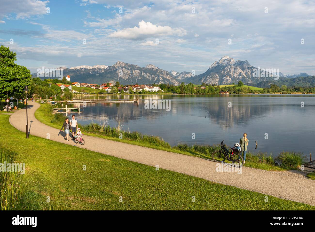 La gente cammina sulla riva del lago Hopfen di fronte alle montagne delle Alpi Ammergau al sole del pomeriggio, Allgäu, Baviera, Germania Foto Stock