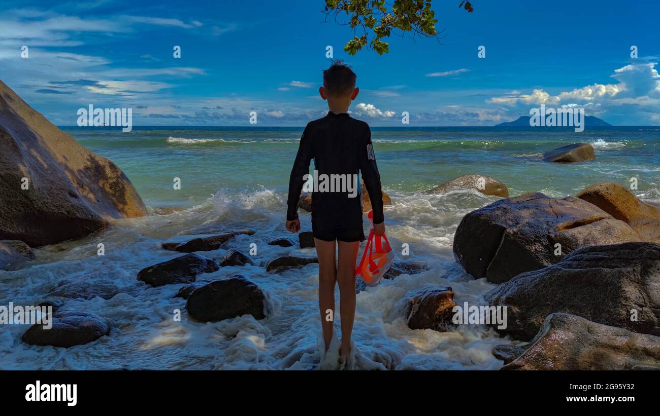 Vista posteriore sul ragazzo in piedi sulla riva del mare in una maschera di tenuta costume da bagno Foto Stock