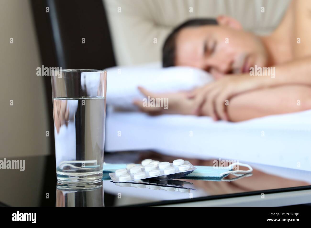 Uomo malato che dorme in un letto, pillole e bicchiere d'acqua in primo piano. Concetto di malattia, febbre, sintomi del coronavirus Foto Stock