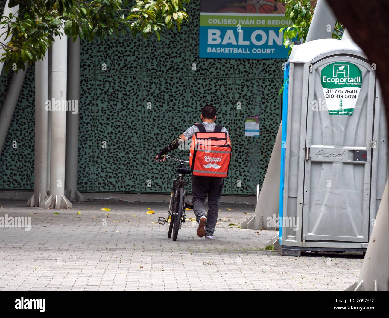 Medellin, Colombia - Luglio 21 2021: Latin Men cammina accanto alla sua bicicletta con 'Rappi' zaino in un parco con un segno che dice : 'Balon' Foto Stock