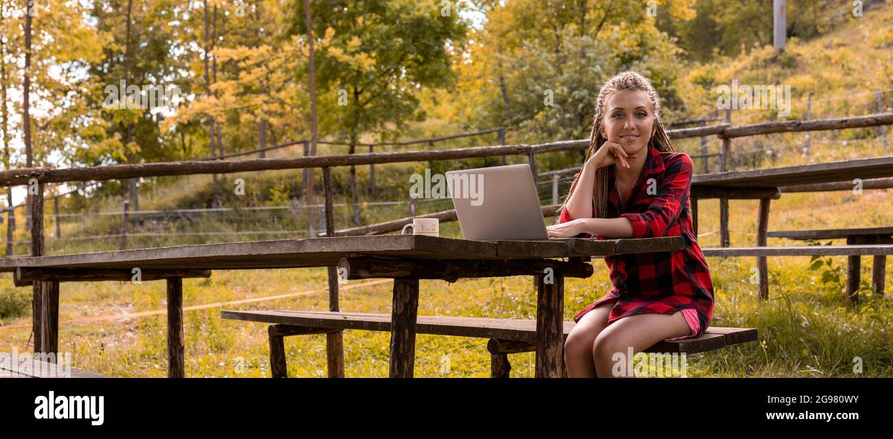 ritratto di una giovane donna attraente con acconciatura intrecciata e camicia plaid, interrompe il suo lavoro al laptop per sorridere a voi in attesa Foto Stock