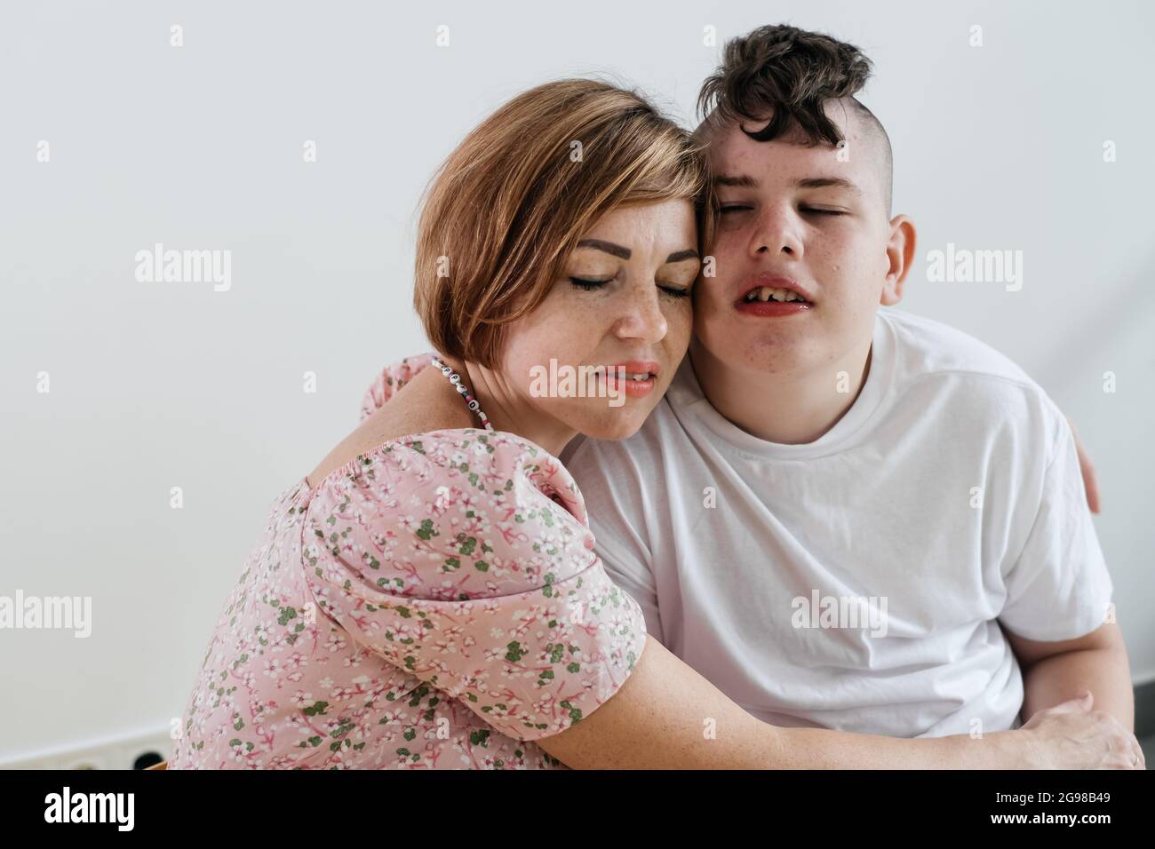 Madre abbracciando disabile ragazzo 17 anni in sedia a rotelle con paralisi cerebrale su sfondo bianco. Donna assiste la persona con bisogni speciali. Emotivo Foto Stock