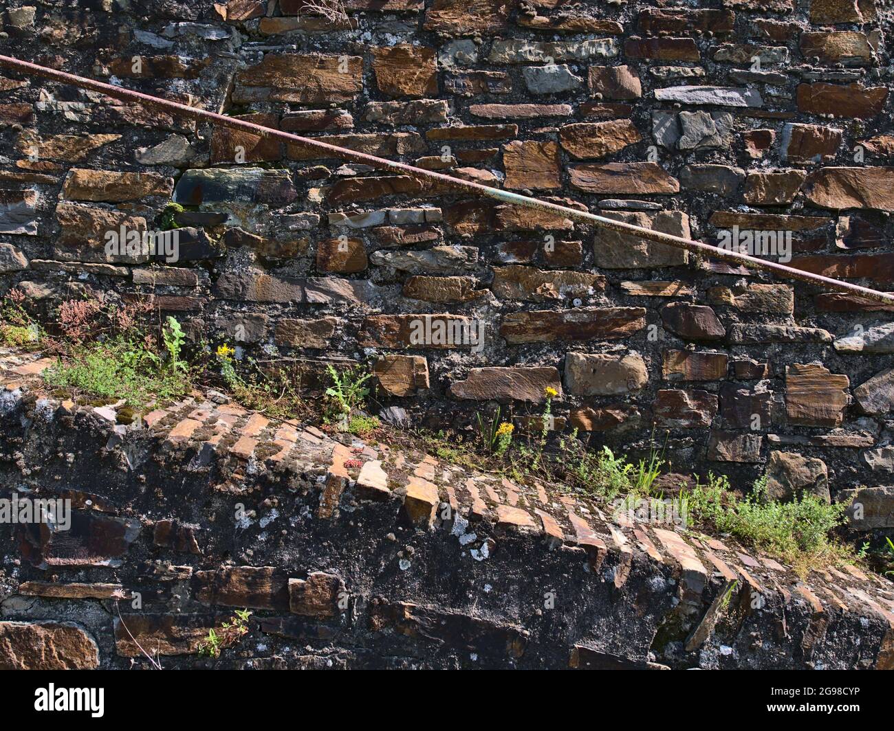 Vista frontale della parete in pietra testurizzata con percorso ascendente e rusty ringhiera in metallo con mattoni a motivi arancioni e erbacce tra vicino a Boppard, Germania. Foto Stock