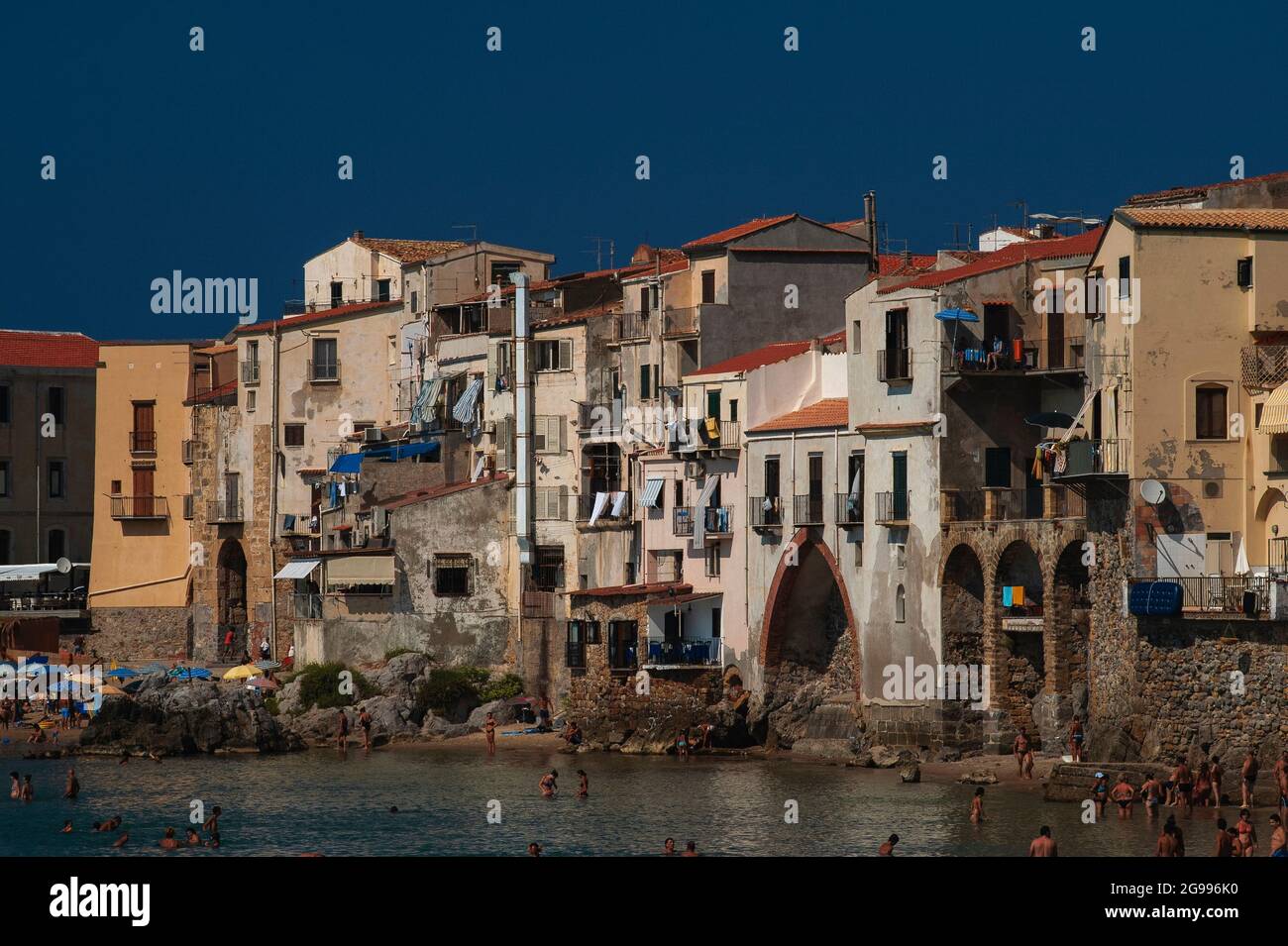 I bagnanti bronzati dal sole si trovano a pale in arrache del Mar Tirreno sotto le mura ocre e bianche delle case del centro storico di Cefalù, Sicilia, Italia, costruite sulle macerie ad arco resti delle mura medievali della città, progettate per proteggere la città costiera settentrionale da attacchi di mare e incursioni piratesche. Cefalù, fondata dagli antichi Greci e occupata a sua volta da Romani, Bizantini, Arabi e Normanni, è diventata una delle località turistiche più famose della Sicilia. Foto Stock