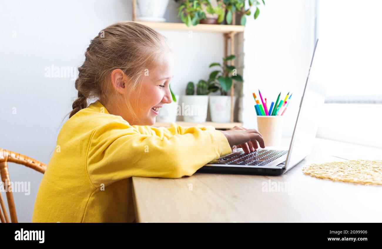 Caucasica ragazza allegra scuola, digitando su una tastiera portatile mentre si siede vicino alla finestra a casa, blogging, chattare, visualizzare le pagine dei social media o. Foto Stock