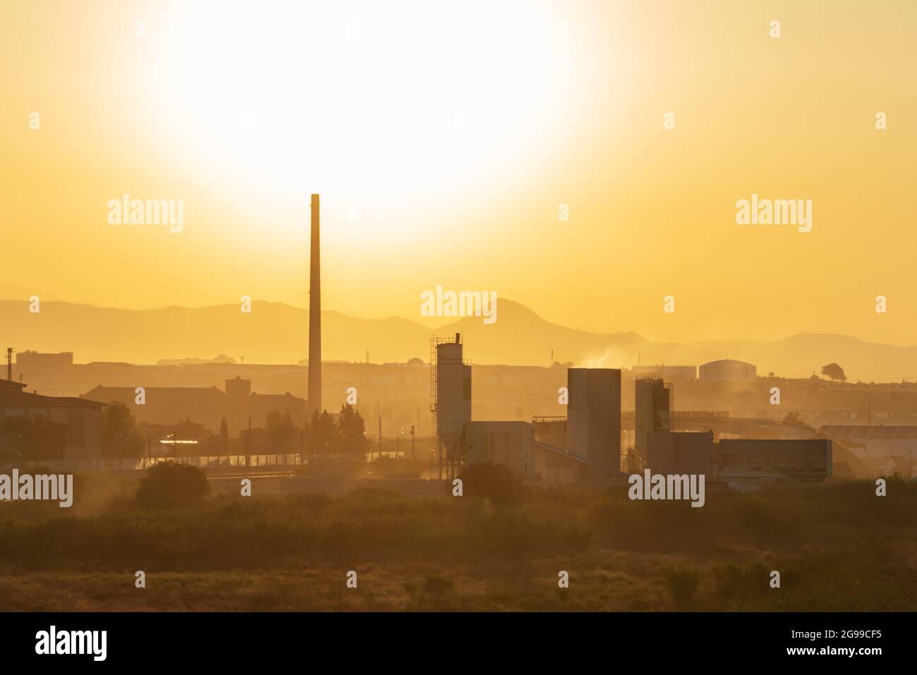 Paesaggio con sfondo industriale, fabbriche, camini, magazzini; illuminato da un grande sole, creando un'atmosfera di alta temperatura ambiente. Foto Stock