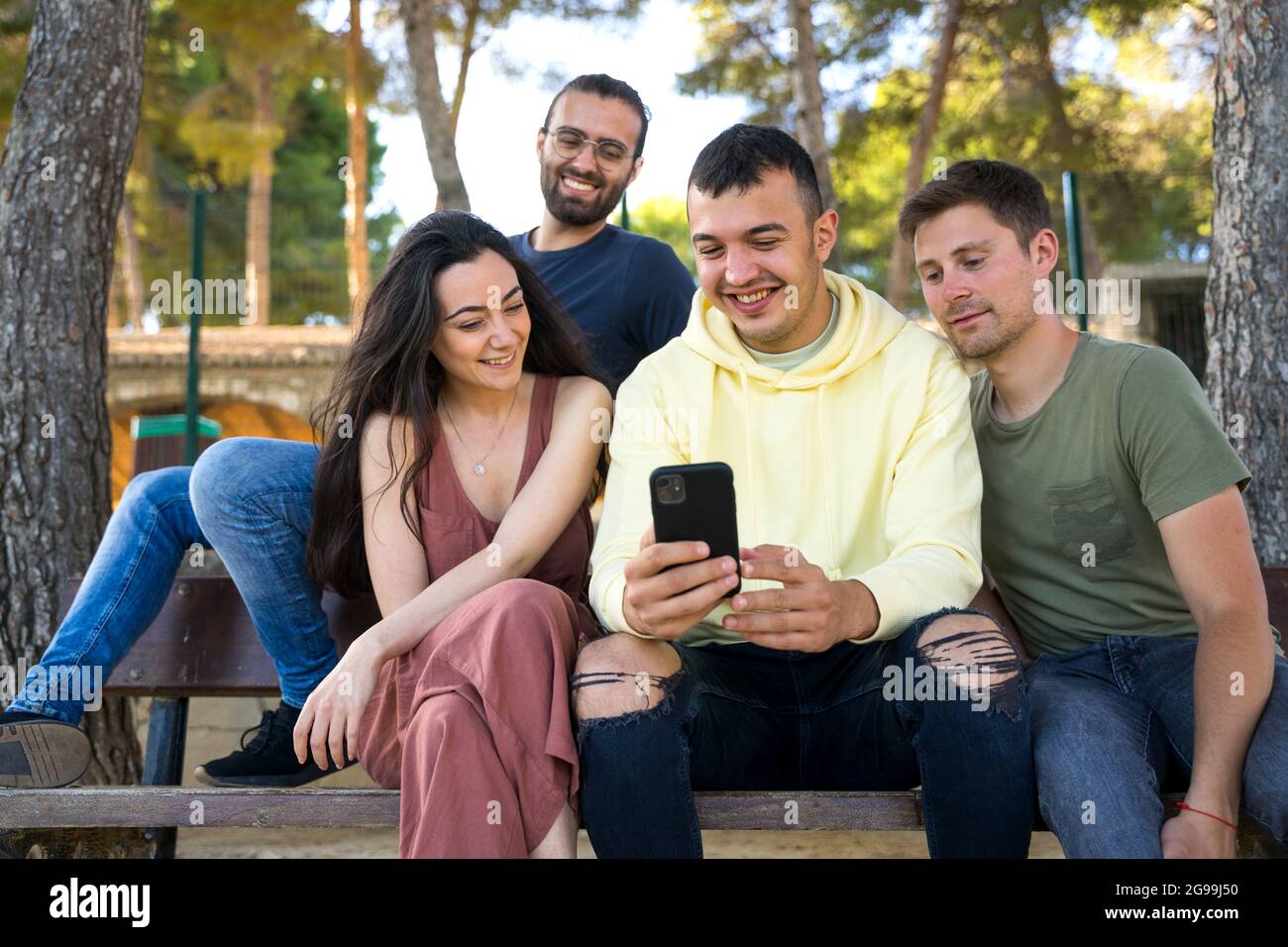 Gruppo di amici seduti su una panca del parco. Sorridono mentre guardano i loro telefoni cellulari. Sono felici e indossano abiti estivi. In un parco Foto Stock