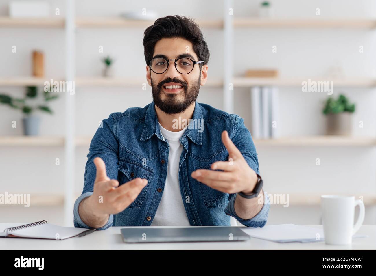 Ritratto di un uomo arabo positivo seduto in ufficio a casa, che parla con la fotocamera, un uomo allegro consulente che condivide idee di business Foto Stock