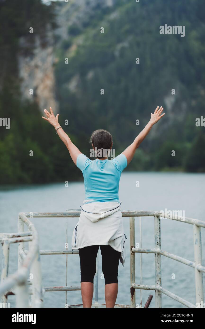 Donna matura in piedi e alzando le braccia verso un lago blu con grandi montagne di fronte. Concetto di viaggio e libertà Foto Stock