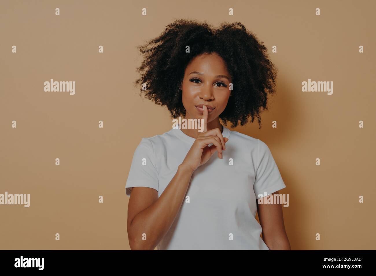 Ritratto in primo piano di una giovane donna afro americana con capelli ondulati in una t-shirt bianca che mostra il segno shh Foto Stock