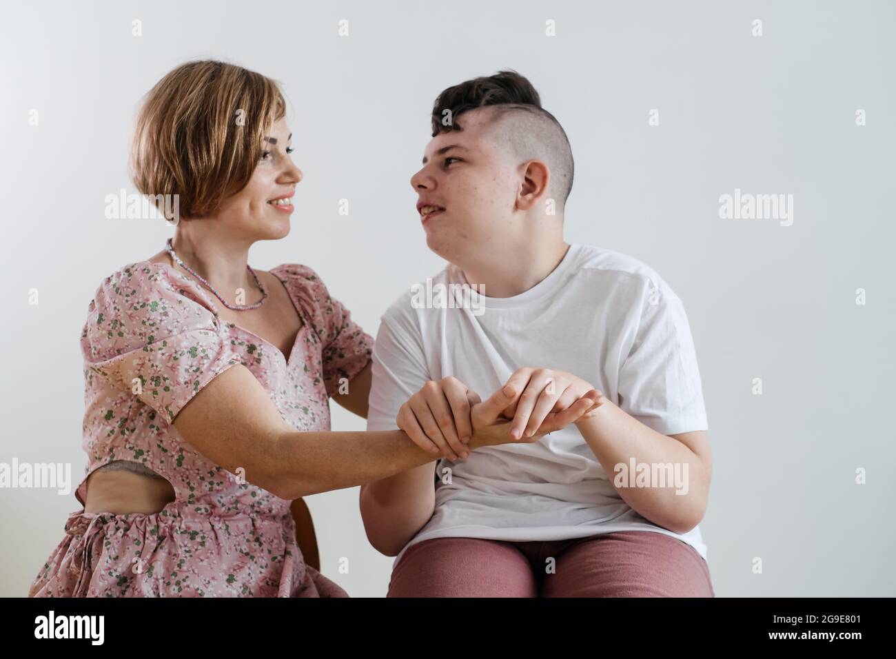 Madre abbracciando disabile ragazzo 17 anni in sedia a rotelle con paralisi cerebrale su sfondo bianco. Donna assiste la persona con bisogni speciali. Emotivo Foto Stock