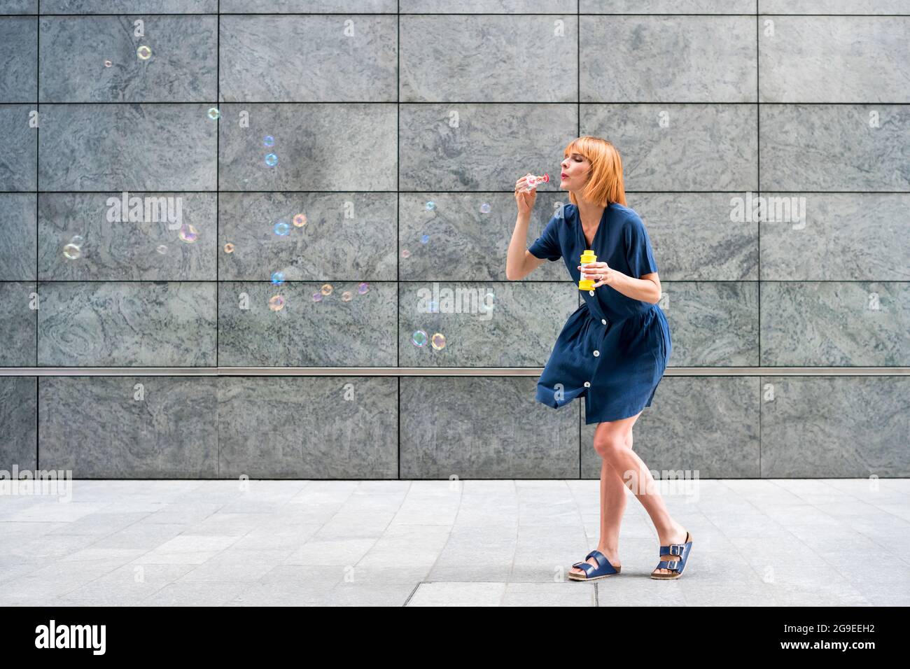 Donna spensierata redhead che celebra bolle di sapone soffiante in città contro una parete grigia di un edificio commerciale in un giorno ventilato con copyspace Foto Stock