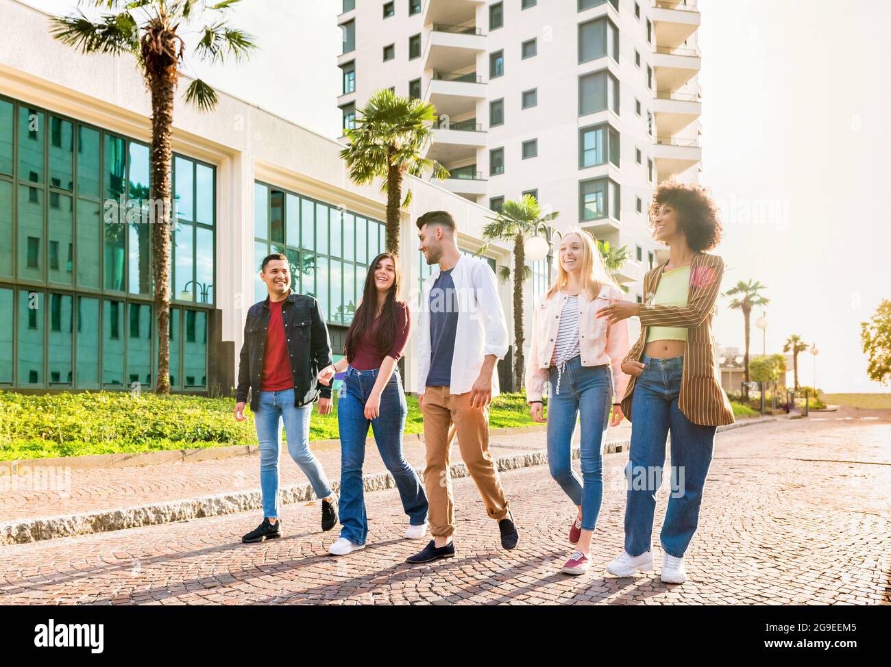 Gruppo multiculturale variegato di giovani amici che camminano in fila lungo una strada urbana chiacchierando e ridendo retroilluminati dal sole in una vista ad angolo basso Foto Stock