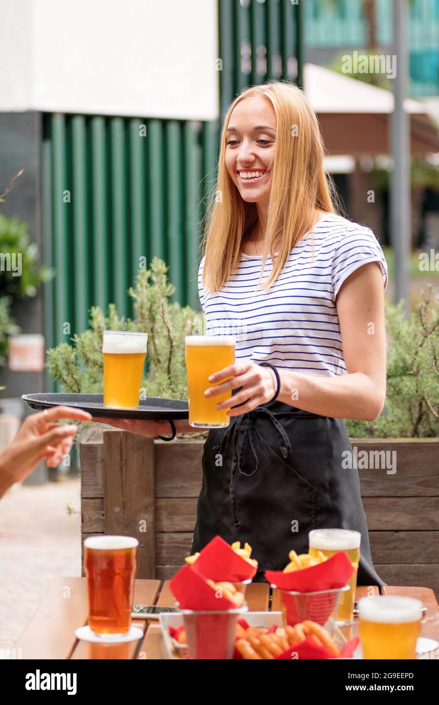 Una cameriera felice e cordiale che serve pinte di birra o birra in un ristorante all'aperto sorridendo mentre il cliente stende una mano per prendere il bicchiere Foto Stock