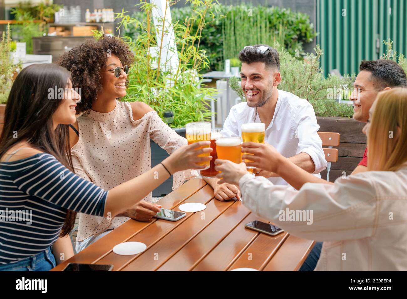 Gruppo di diversi giovani amici che festeggiano in un ristorante all'aperto o in un pub che si tostano a vicenda con birre fredde mentre ridono e sorridono Foto Stock