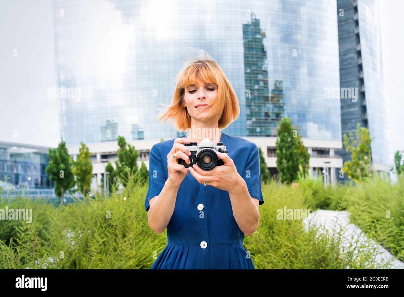 Bella donna rossa scattando foto sulla sua fotocamera in città sullo sfondo di un verde e un moderno blocco torre di vetro in una frizzante giornata estiva Foto Stock