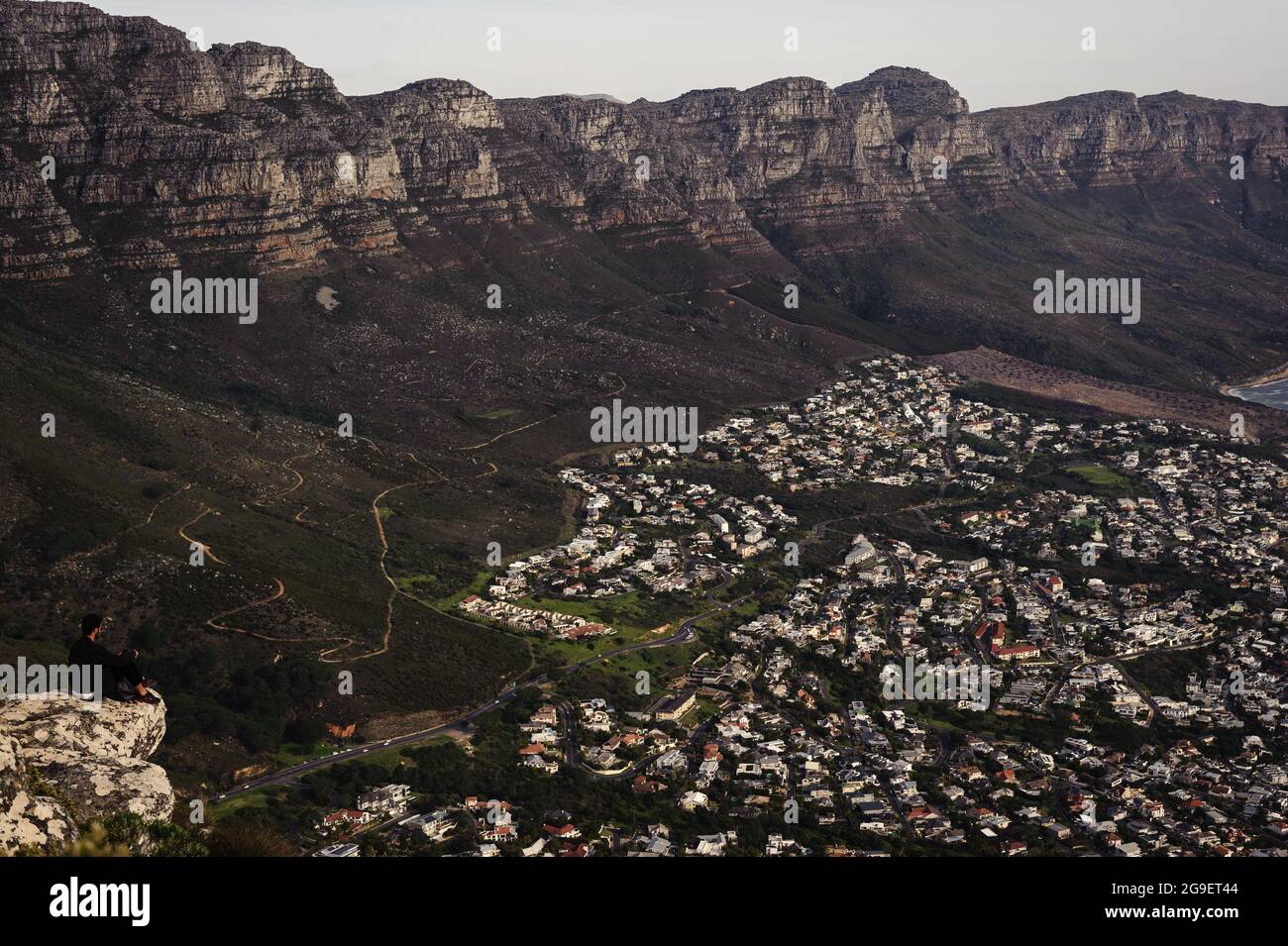 Un uomo salotti sulla montagna Lion's Head di Città del Capo per vedere i 12 Apostoli di Table Mountain e l'elegante sobborgo costiero di Camps Bay sottostante Foto Stock