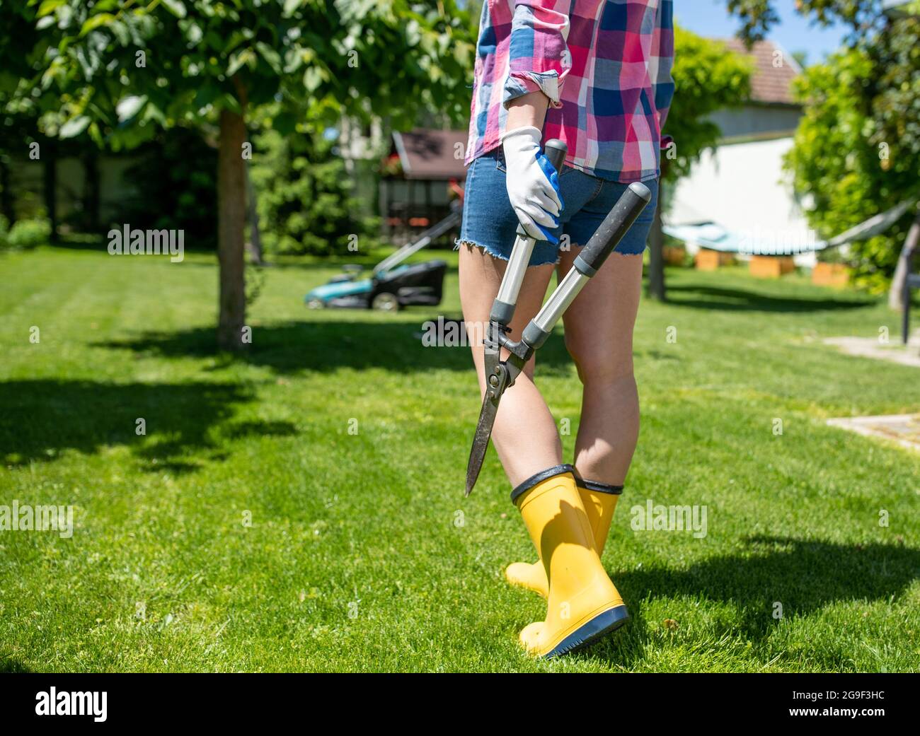 Donna giardiniere che cammina in cantiere tenendo forare cesoie. Vista da dietro la giovane donna che indossa i wellies e che tiene gli attrezzi di giardinaggio. Foto Stock