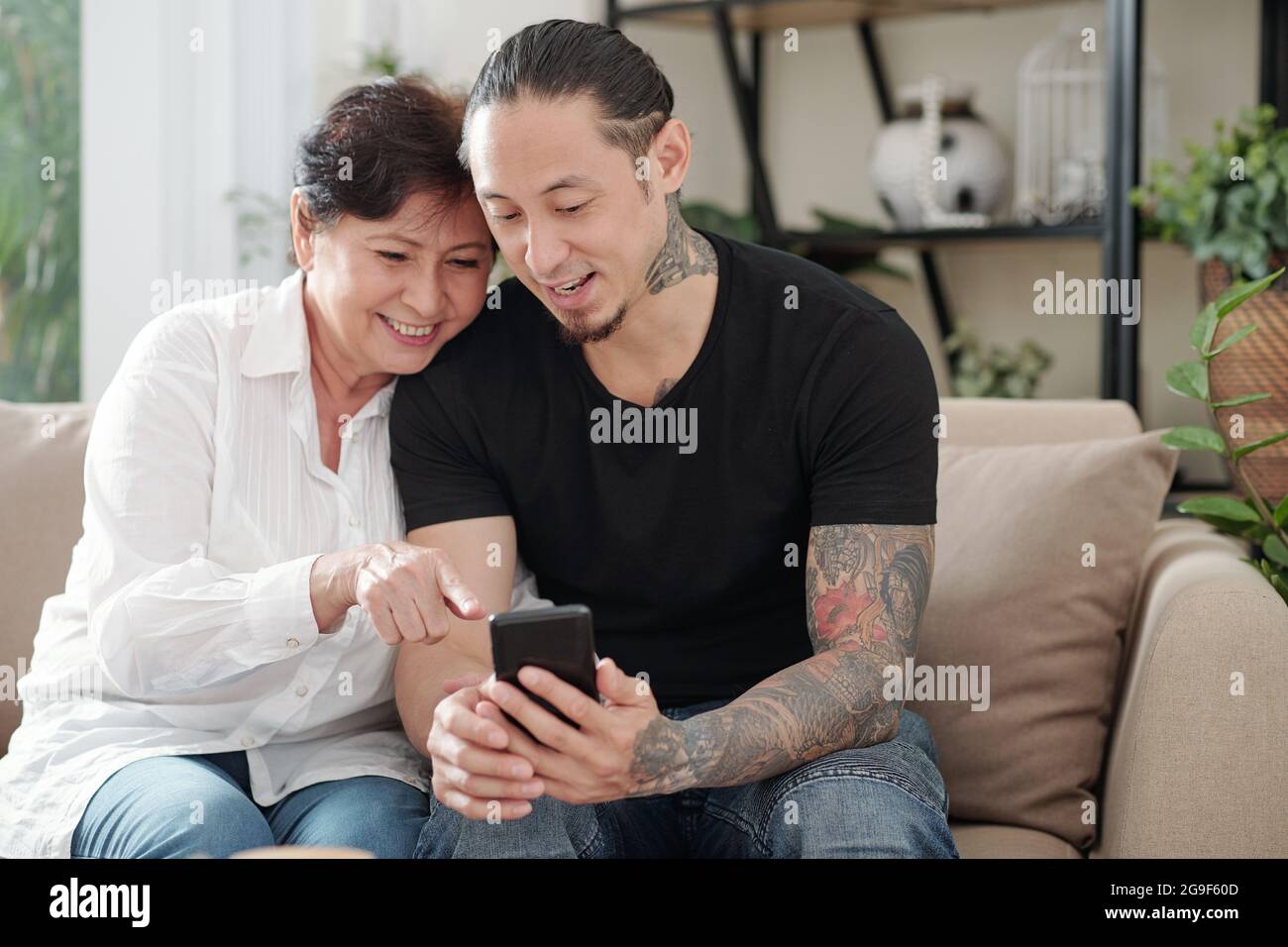 Figlio maggiore che mostra le foto sul suo cellulare alla sua madre felice mentre si riposano sul divano a casa Foto Stock