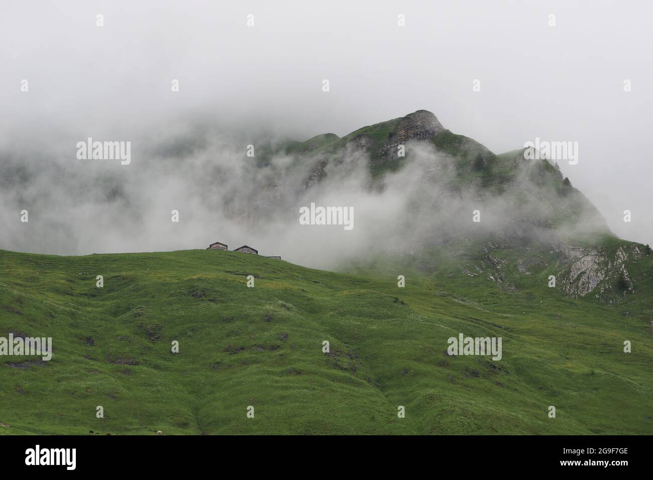 Case coloniche, prato verde e cima di montagna che si allungano dalla nebbia e dalle nuvole. Foto Stock