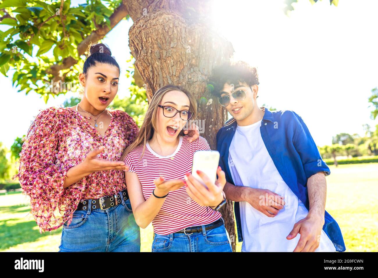 Tre diversi studenti che fanno volti sorpresi che si diffonde bocca e occhi puntando e guardando smartphone passare il tempo nella natura del parco cittadino. Potere di Foto Stock